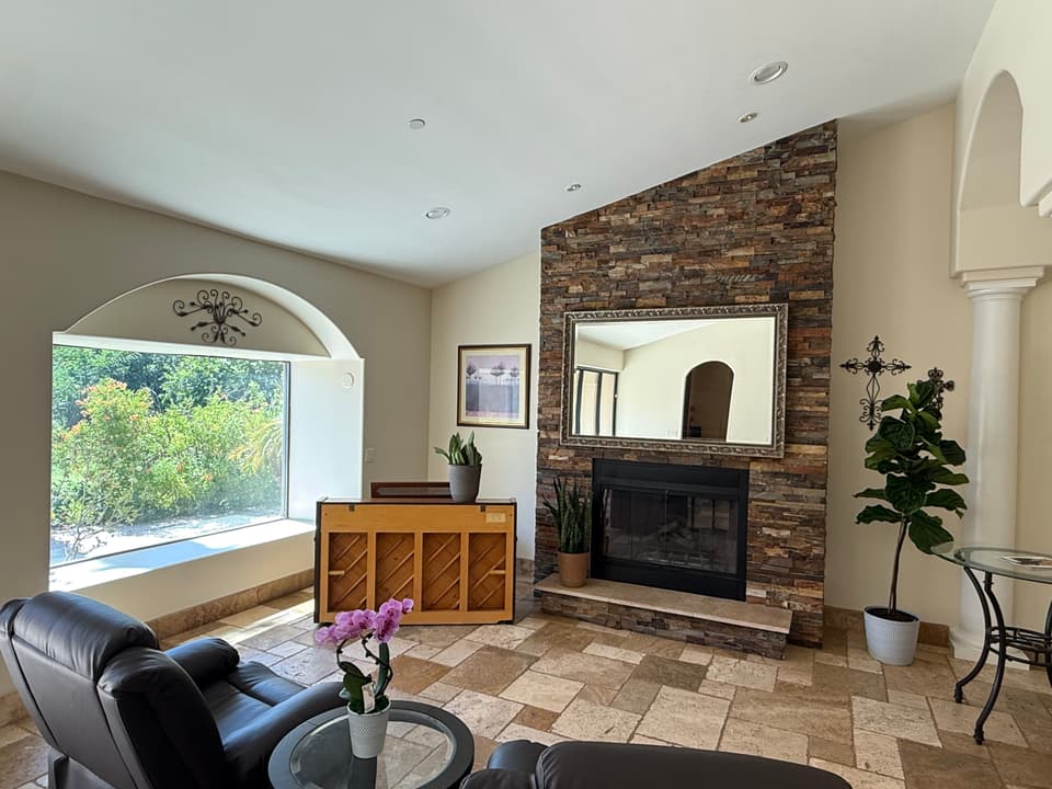 Bright assisted living common area with stone fireplace, large window, comfortable seating, indoor plants, and natural light.
